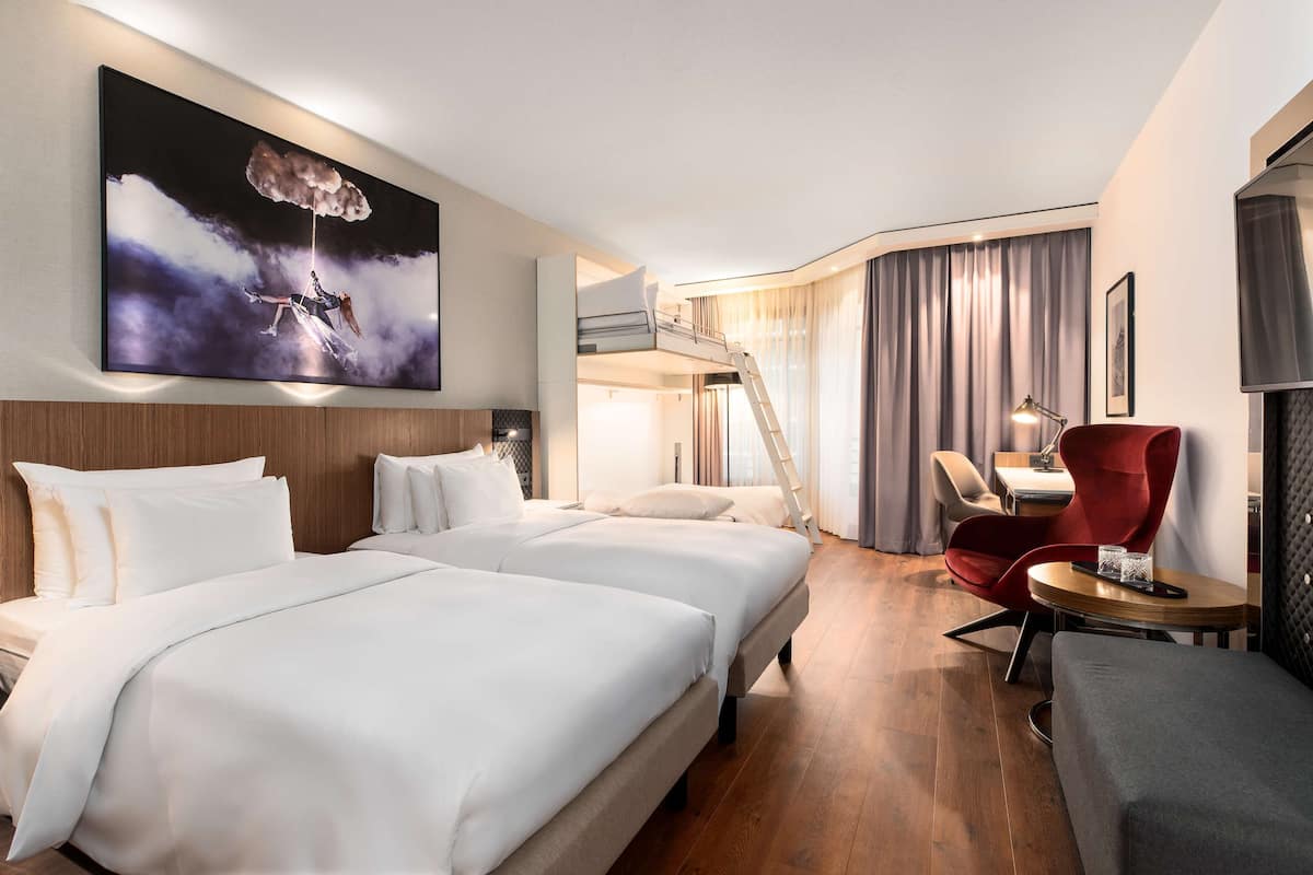 Une chambre d’hôtel avec deux lits, un fauteuil rouge, un bureau et un grand tableau.