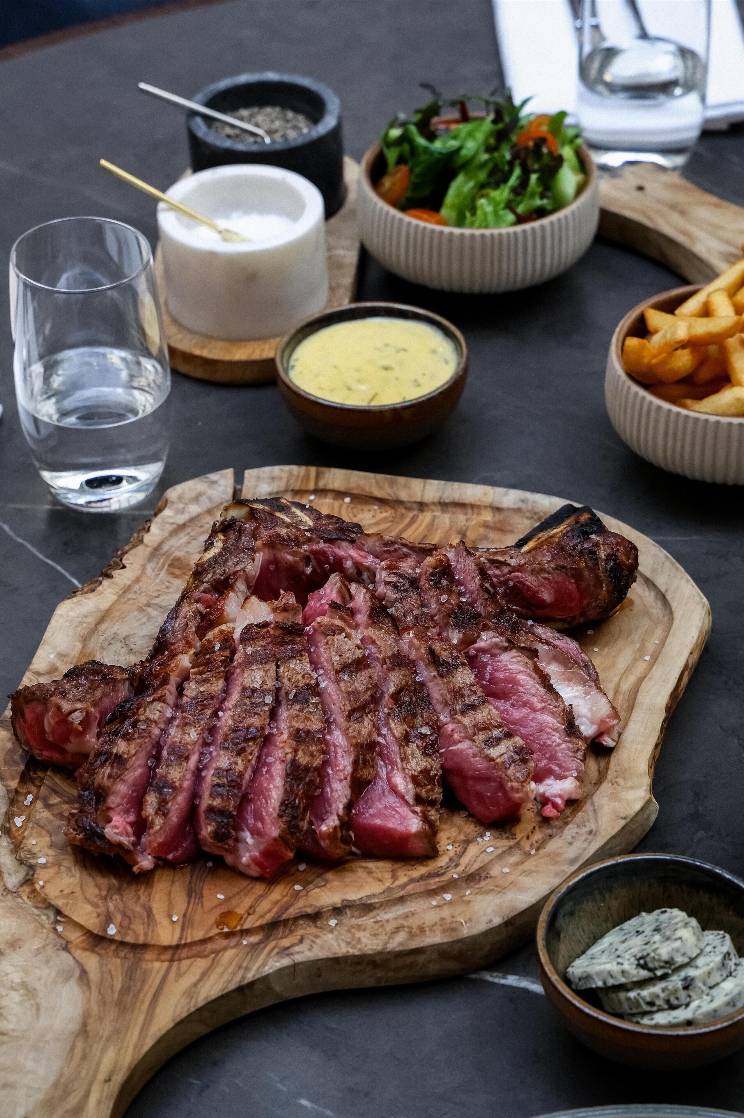 A wooden cutting board with sliced steak, a bowl of salad, and a glass of water on a table.