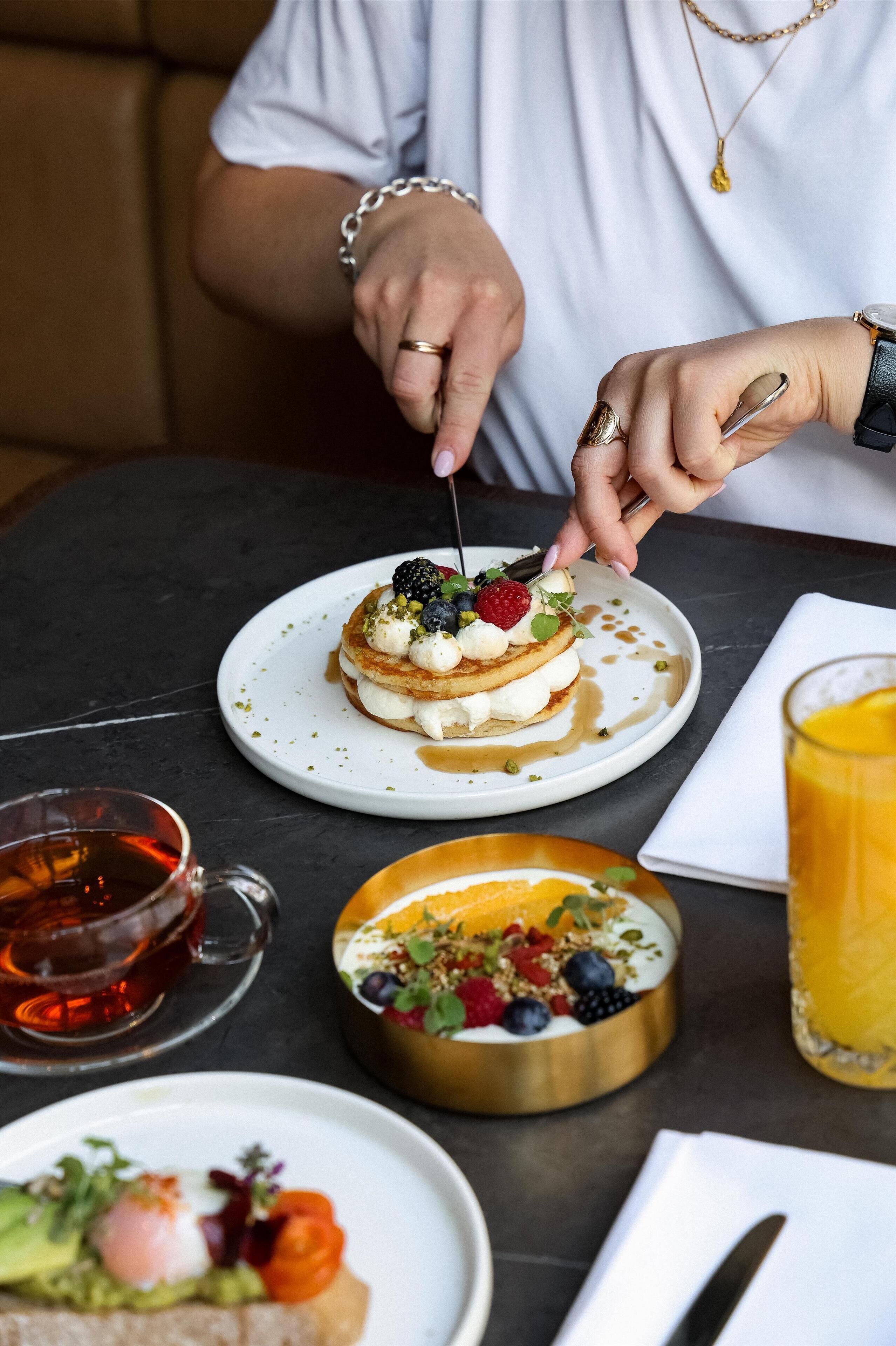 A person cutting a dessert with a fork. There are plates with food, a glass of juice, and a cup of tea on the table.
