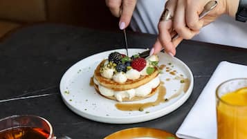 A person cutting a dessert with a fork. There are plates with food, a glass of juice, and a cup of tea on the table.
