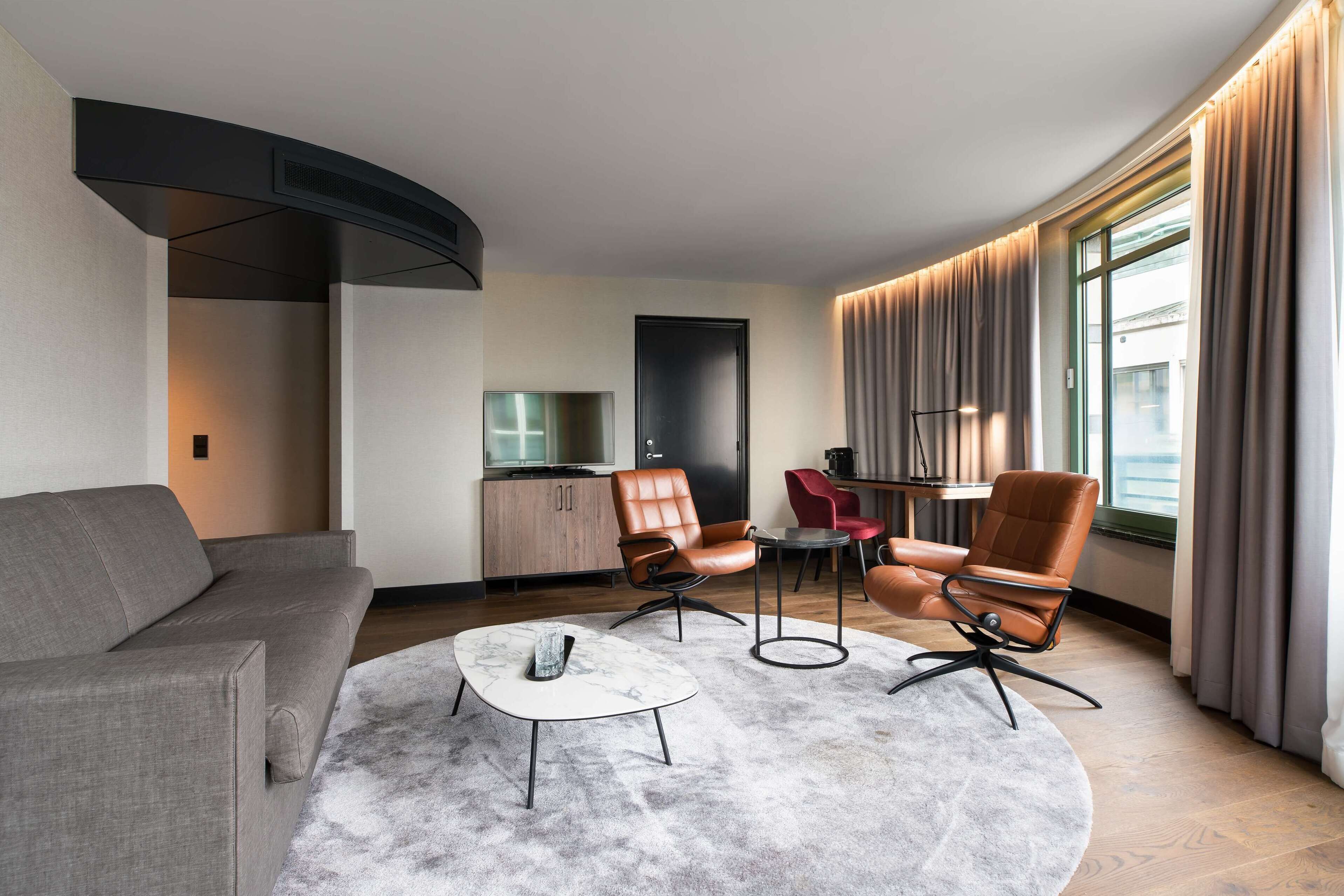 Een moderne woonkamer met een bank, twee leren fauteuils, een salontafel en een eethoek met een tafel en stoelen.
