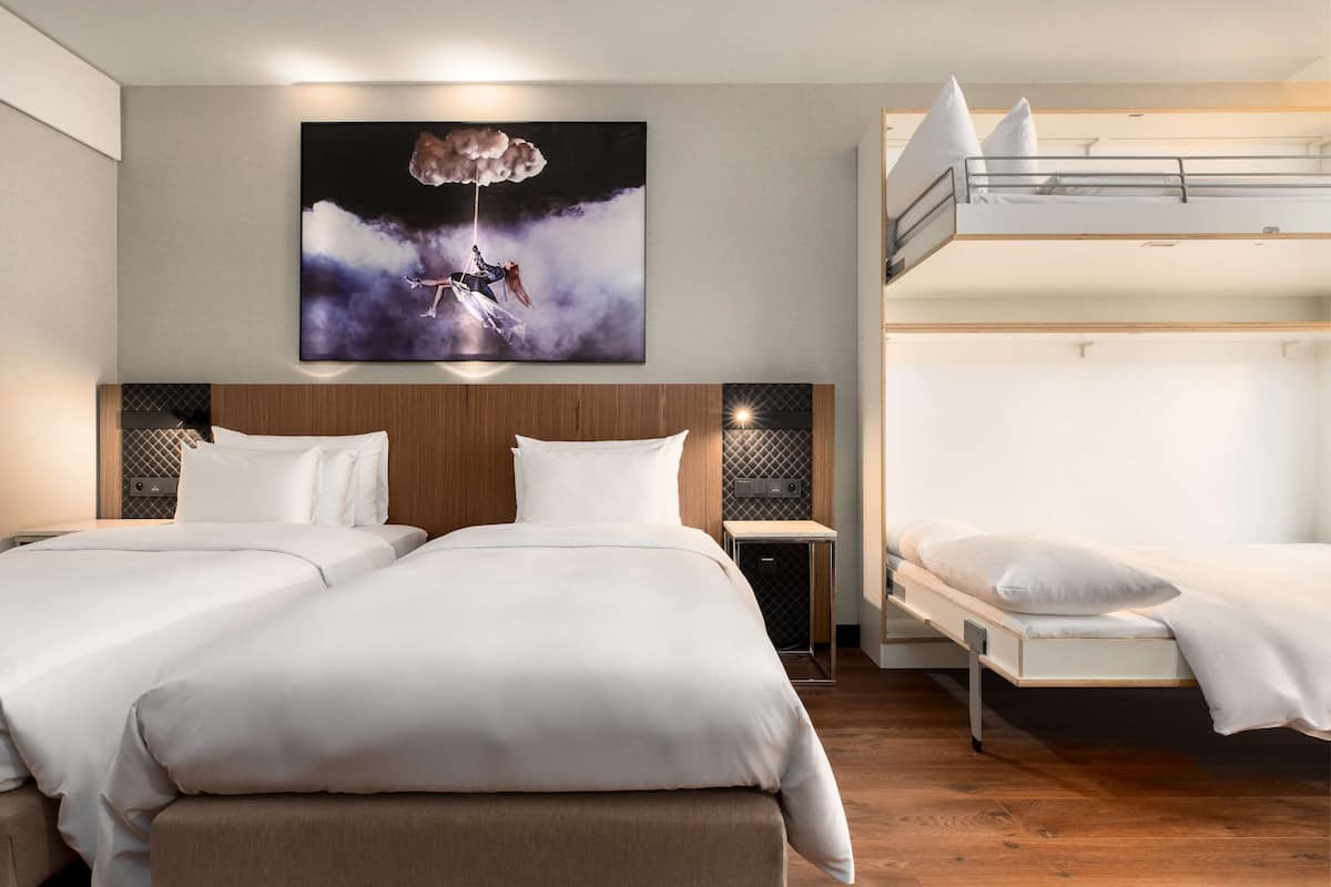 Une chambre d’hôtel avec deux lits, un lit superposé, une tête de lit en bois, une table de chevet, une œuvre d’art accrochée au mur et un plancher en bois.