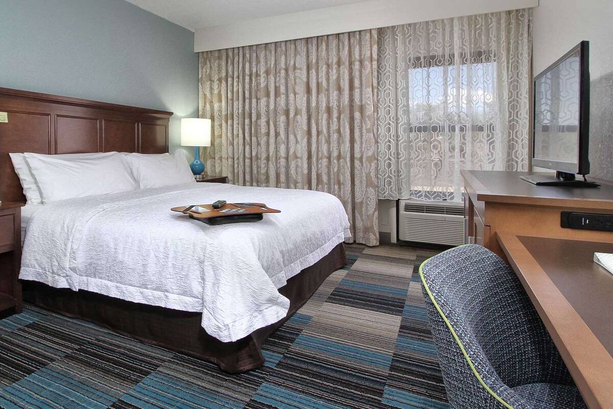 A hotel room with a large bed, a desk with a computer, a chair, a nightstand with a phone, and a window with curtains.