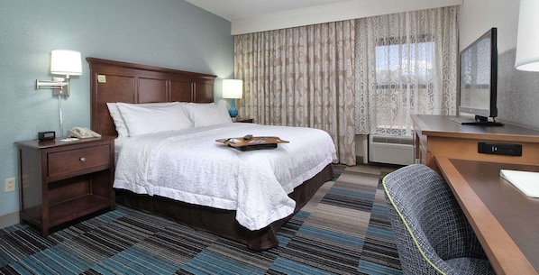 A hotel room with a large bed, a desk with a computer, a chair, a nightstand with a phone, and a window with curtains.