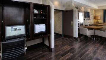 Una habitación de hotel con piso de madera oscura, un escritorio con impresora, una zona de estar con sofá y sillones, y un televisor sobre un mueble.