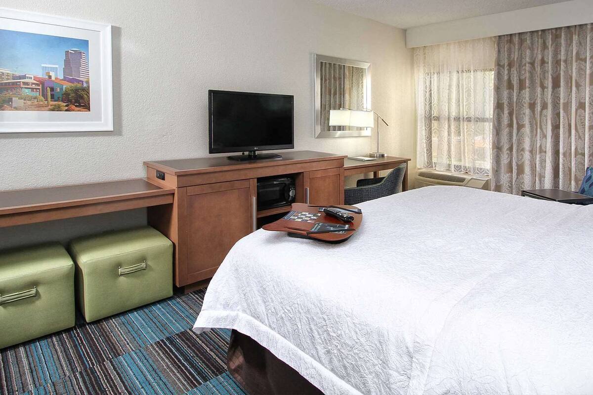 A hotel room with a bed, a desk with a television, a chair, a small table, and a window with curtains.