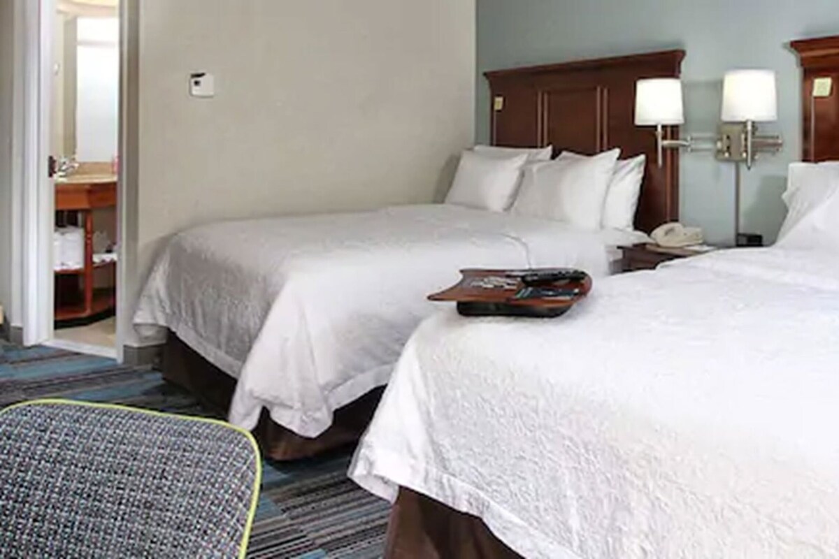 A hotel room with two beds, a nightstand with a lamp, a chair, and a tray with items on the bed.