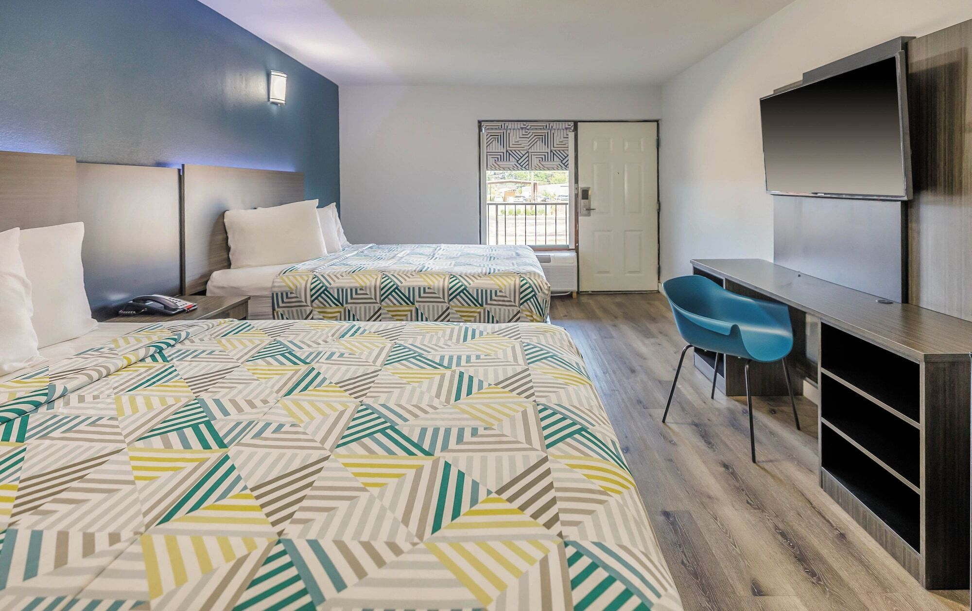 A hotel room with two beds, a desk, a chair, a TV, and a door leading outside.