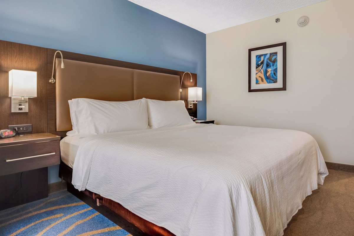 Un cuarto de hotel con una cama grande, dos mesitas de noche, una lámpara de noche, una paleta de colores azul y beige, y un cuadro abstracto enmarcado en la pared.