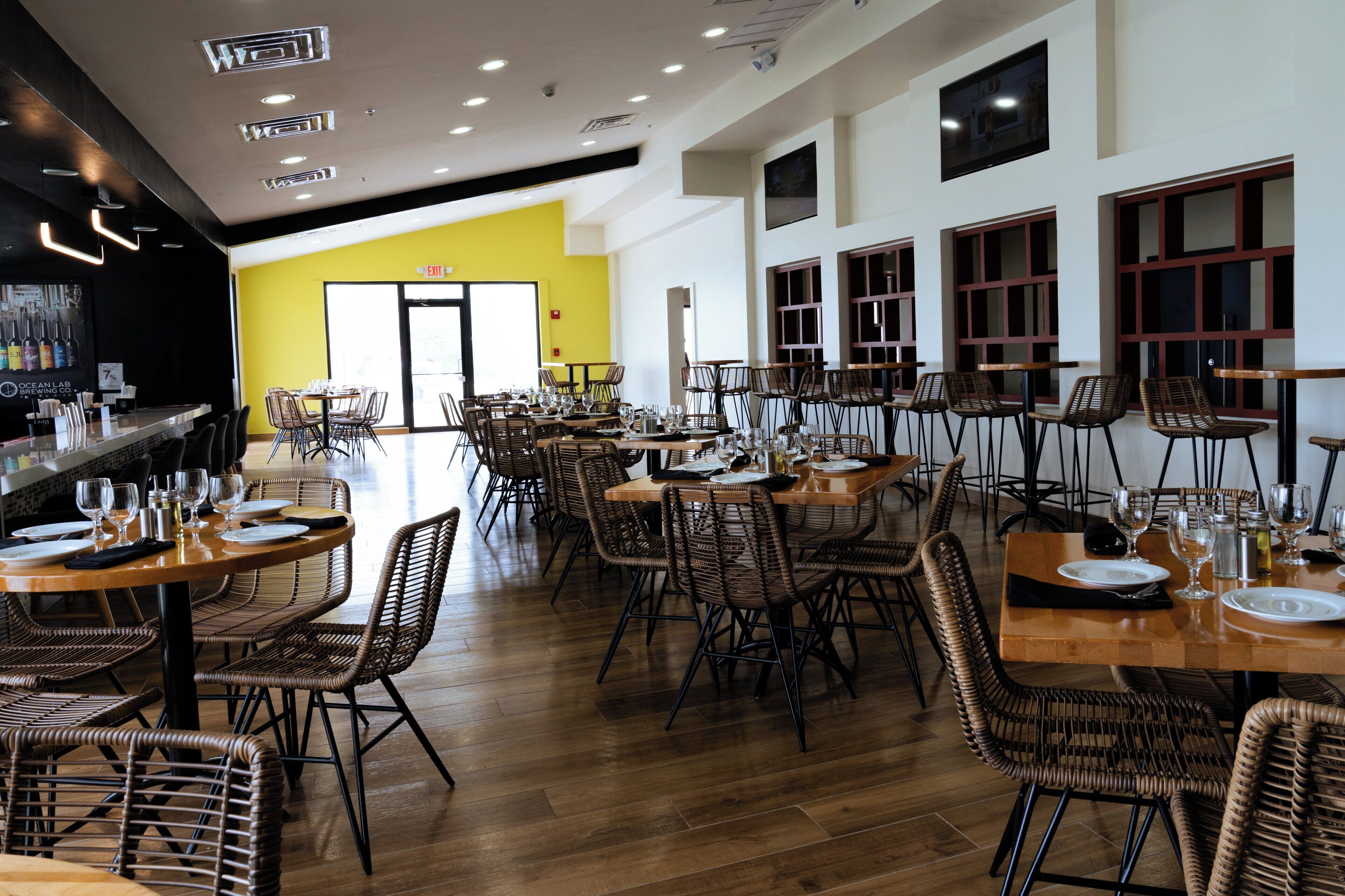 A spacious restaurant with multiple dining tables and chairs, a bar area, and large windows.