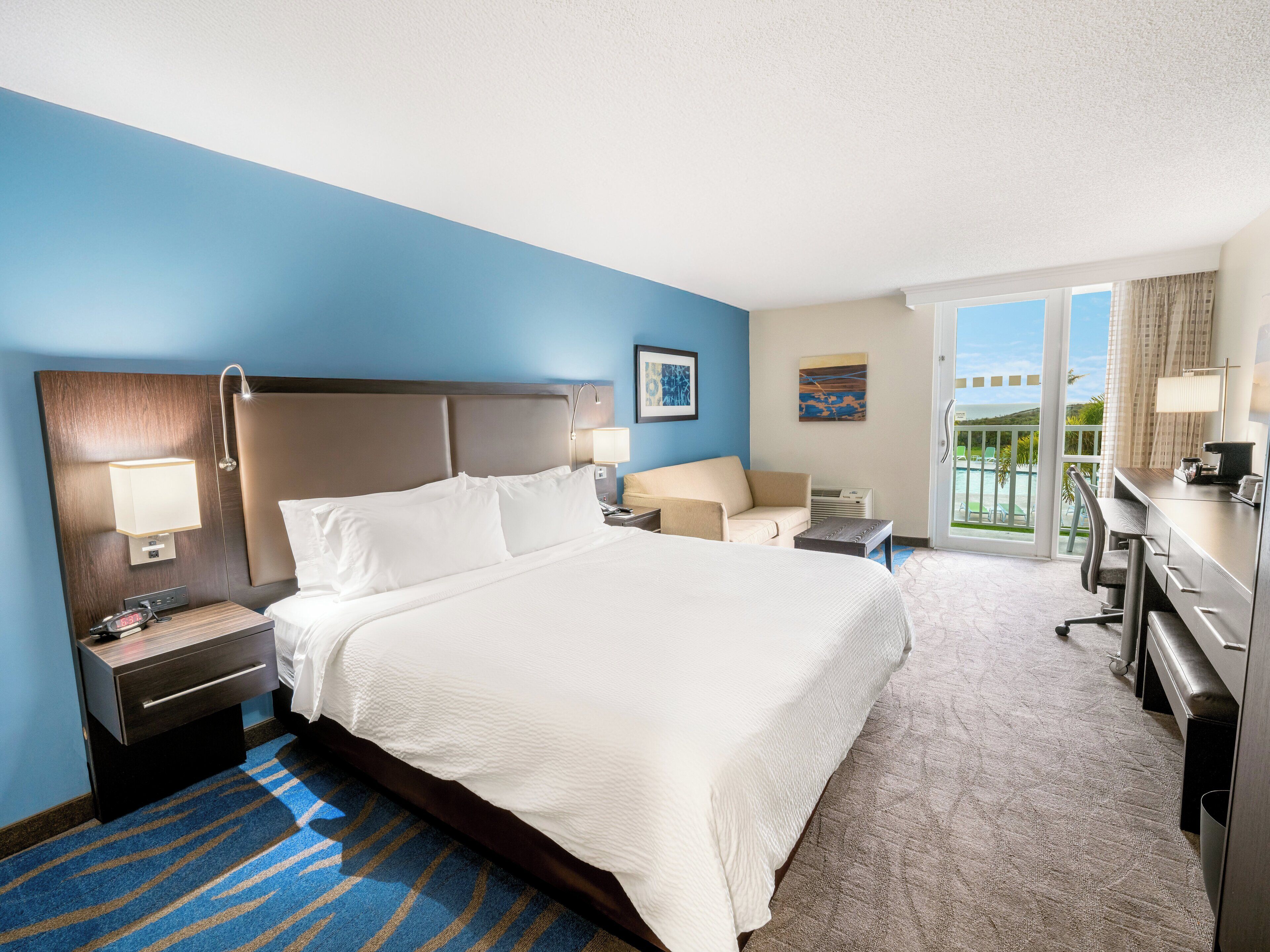 Quarto de hotel com uma cama grande, uma escrivaninha, uma cadeira, uma mesa de cabeceira, uma luminária e vista para o exterior.