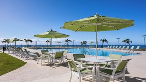 Un espace aménagé au bord de la piscine, avec des chaises longues et des tables, des parasols et une vue sur l’océan.