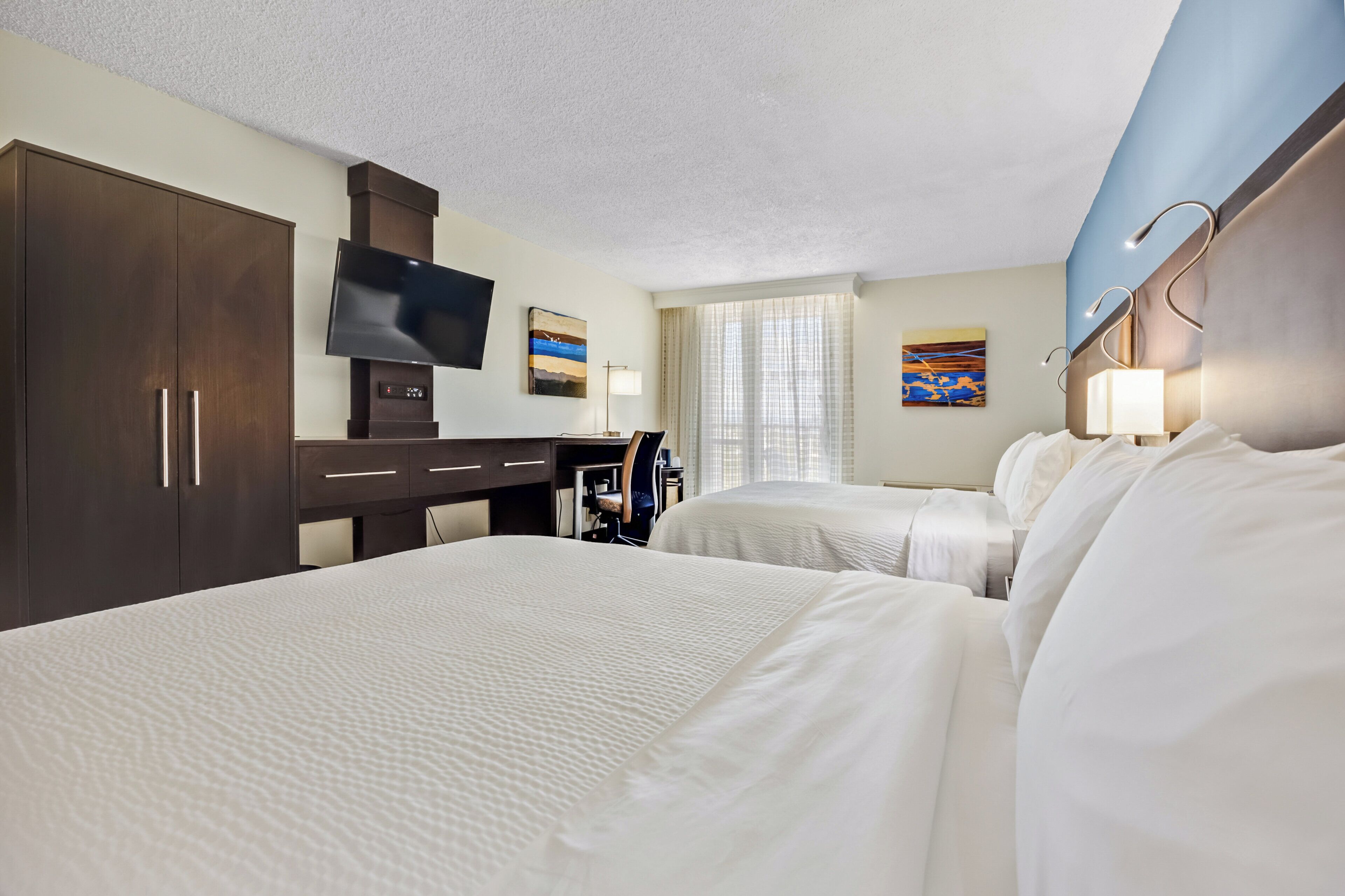 A hotel room with two beds, a desk with a chair, a TV, and artwork on the walls.