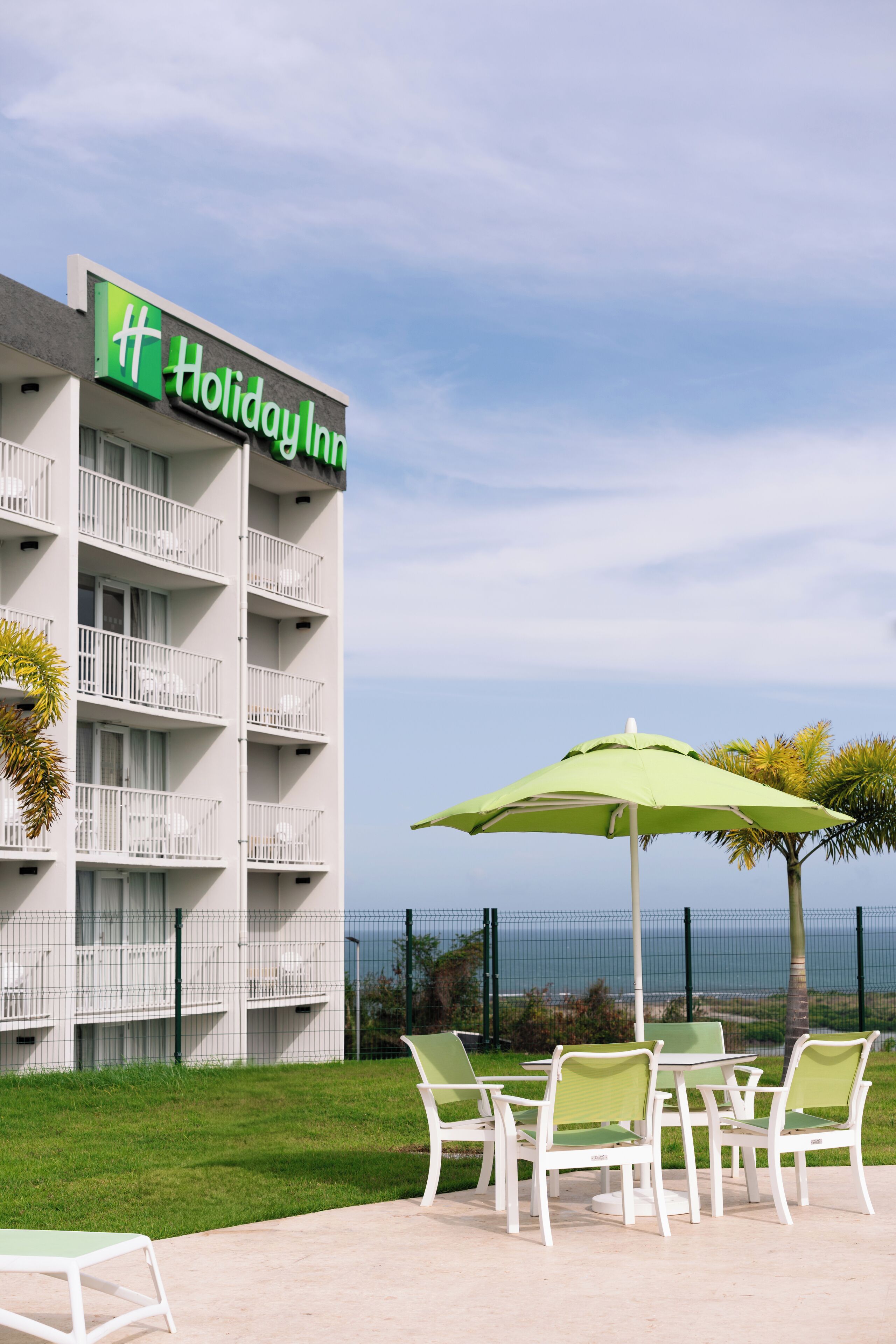 Edificio de Holiday Inn con balcón, zona de estar al aire libre con mesas y sillas, y vista al océano.