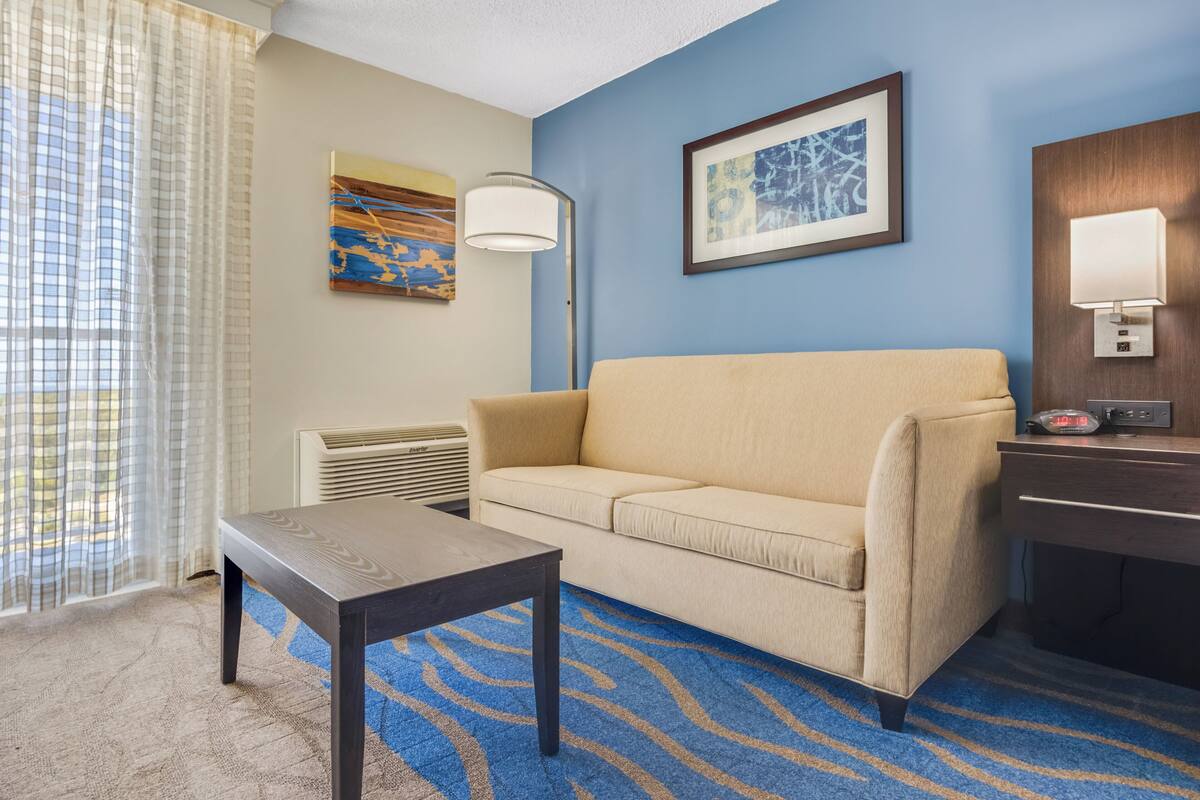 A hotel room with a beige sofa, a small wooden table, a blue patterned carpet, a framed abstract painting, a floor lamp, and a bedside table with a lamp.