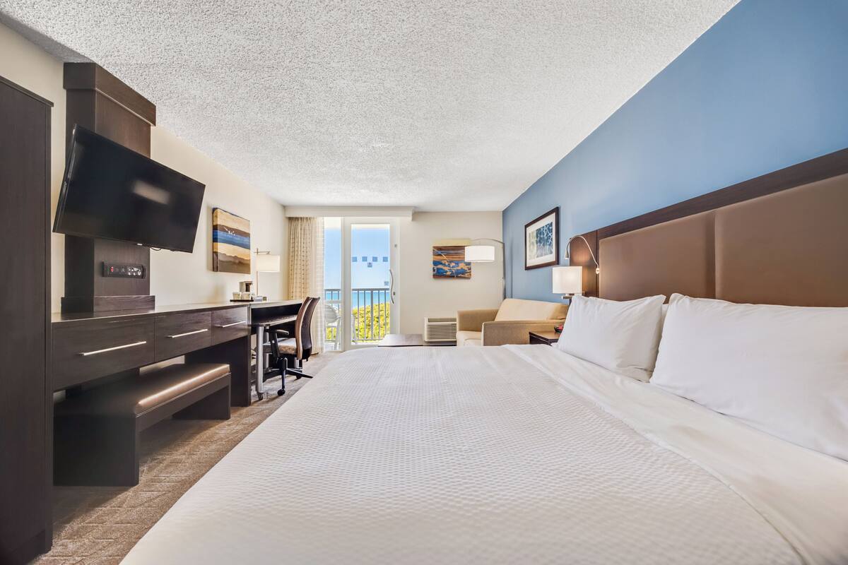 A hotel room with a large bed, a desk with a flat-screen TV, a chair, and a balcony with a view.