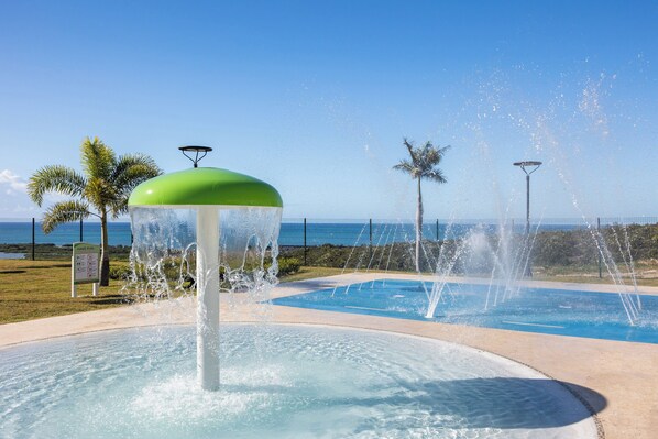 Una fuente de agua con parte superior verde, rodeada por una zona de piscina con palmeras y un cielo despejado.