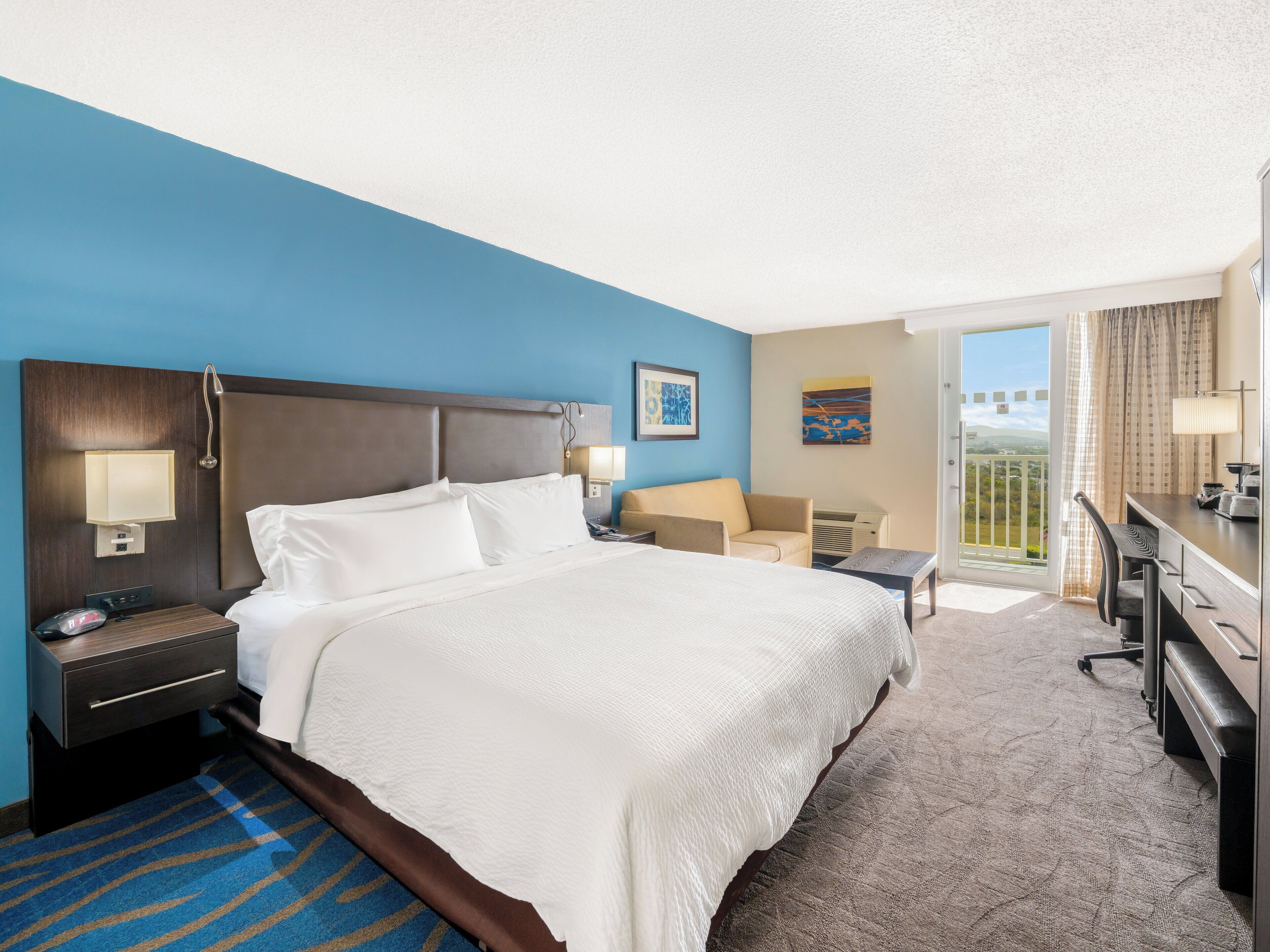 A hotel room with a large bed, a desk, a chair, a small table, and a balcony with a view.
