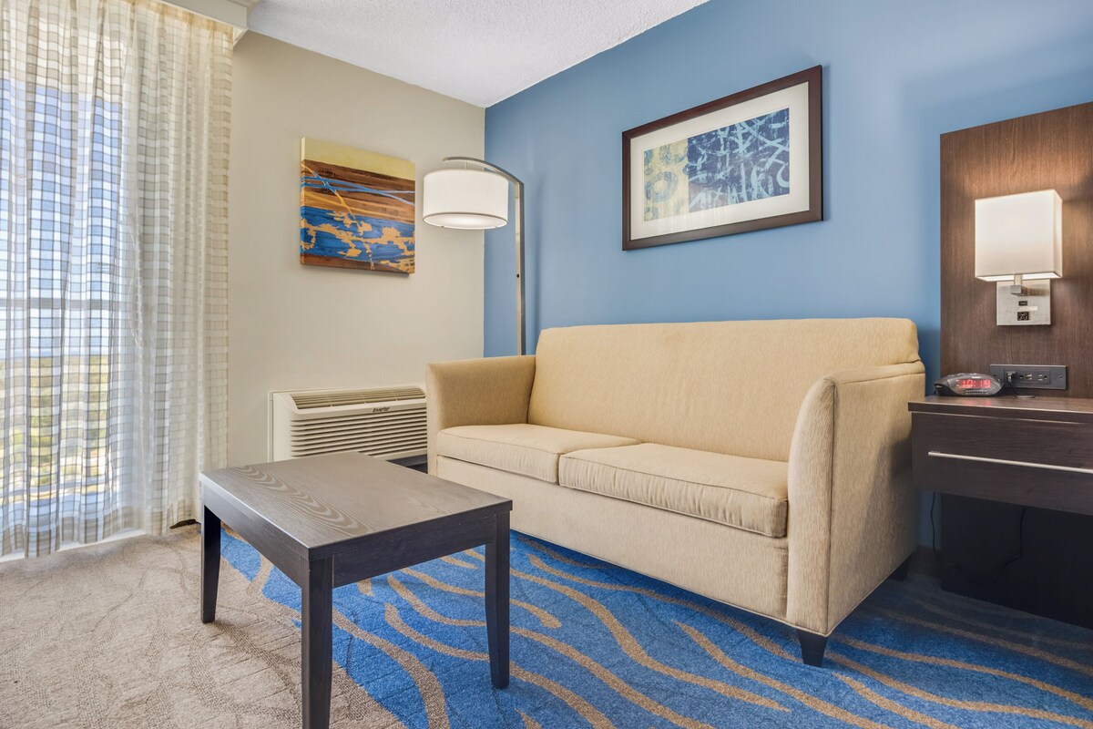 Una habitación de hotel con un sofá beige, una pequeña mesa de madera, una alfombra azul estampada, un cuadro abstracto enmarcado, una lámpara de pie y una mesita de noche con lámpara.