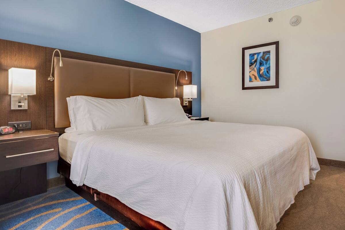 A hotel room with a large bed, two bedside tables, a nightstand, a blue and beige color scheme, and a framed abstract painting on the wall.