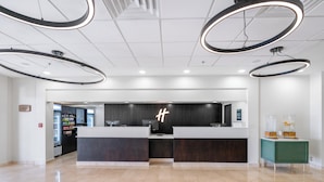 Un espace d’accueil moderne, au design épuré et minimaliste, comprenant un comptoir d’accueil, un distributeur automatique et une petite table d’appoint verte agrémentée d’objets décoratifs.