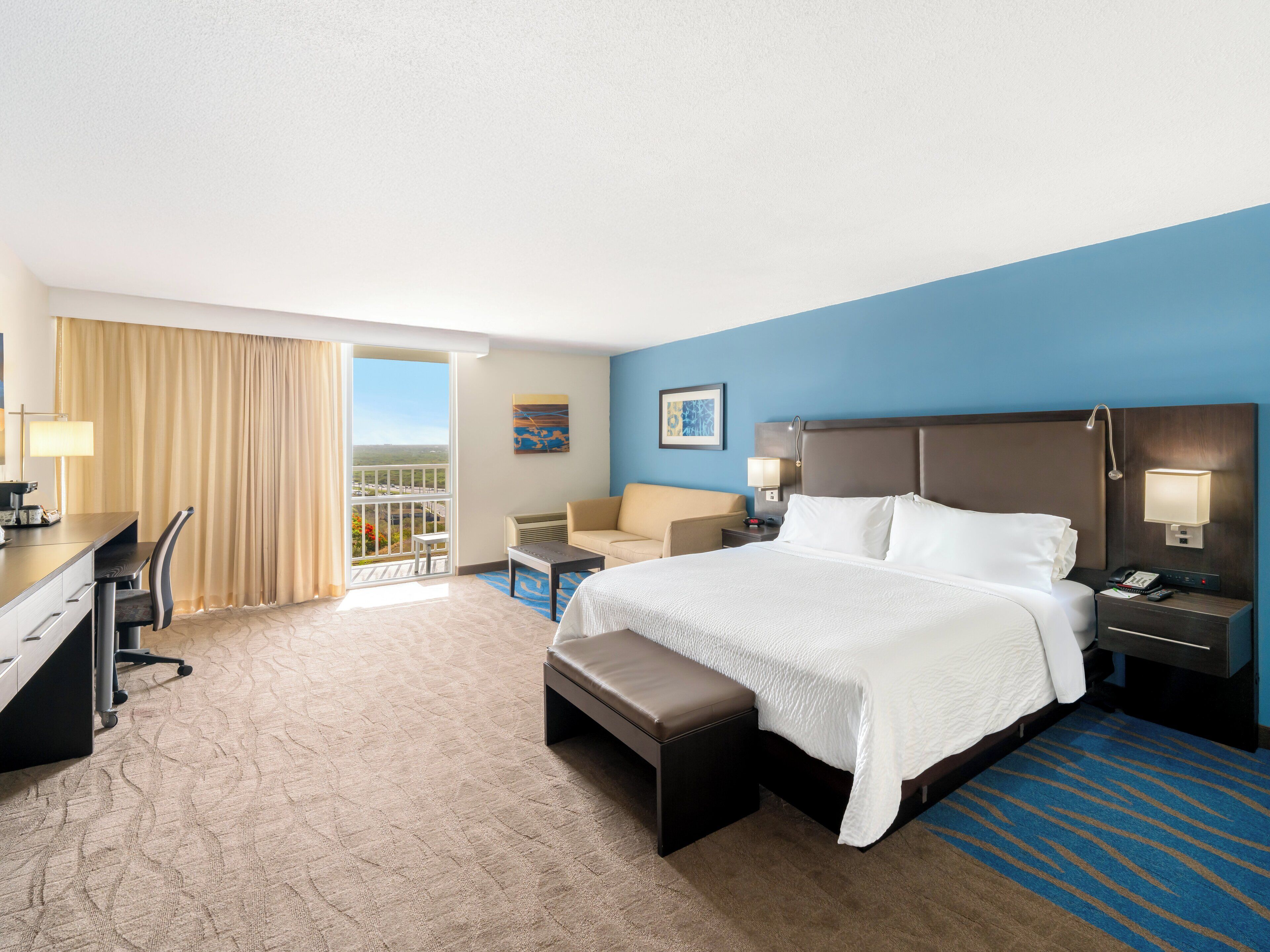 A hotel room with a large bed, a desk, a chair, a sofa, and a balcony with a view.