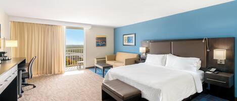 A hotel room with a large bed, a desk, a chair, a sofa, and a balcony with a view.