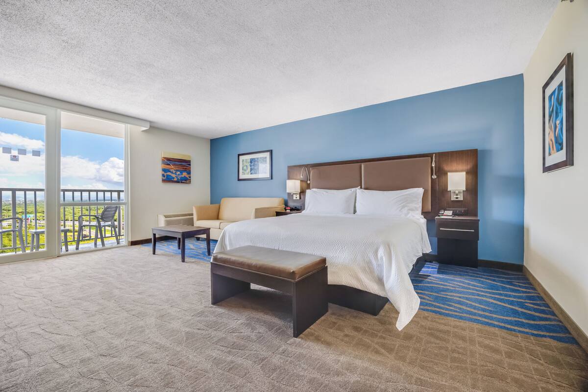 A hotel room with a large bed, a bench, a sofa, and a balcony with chairs.