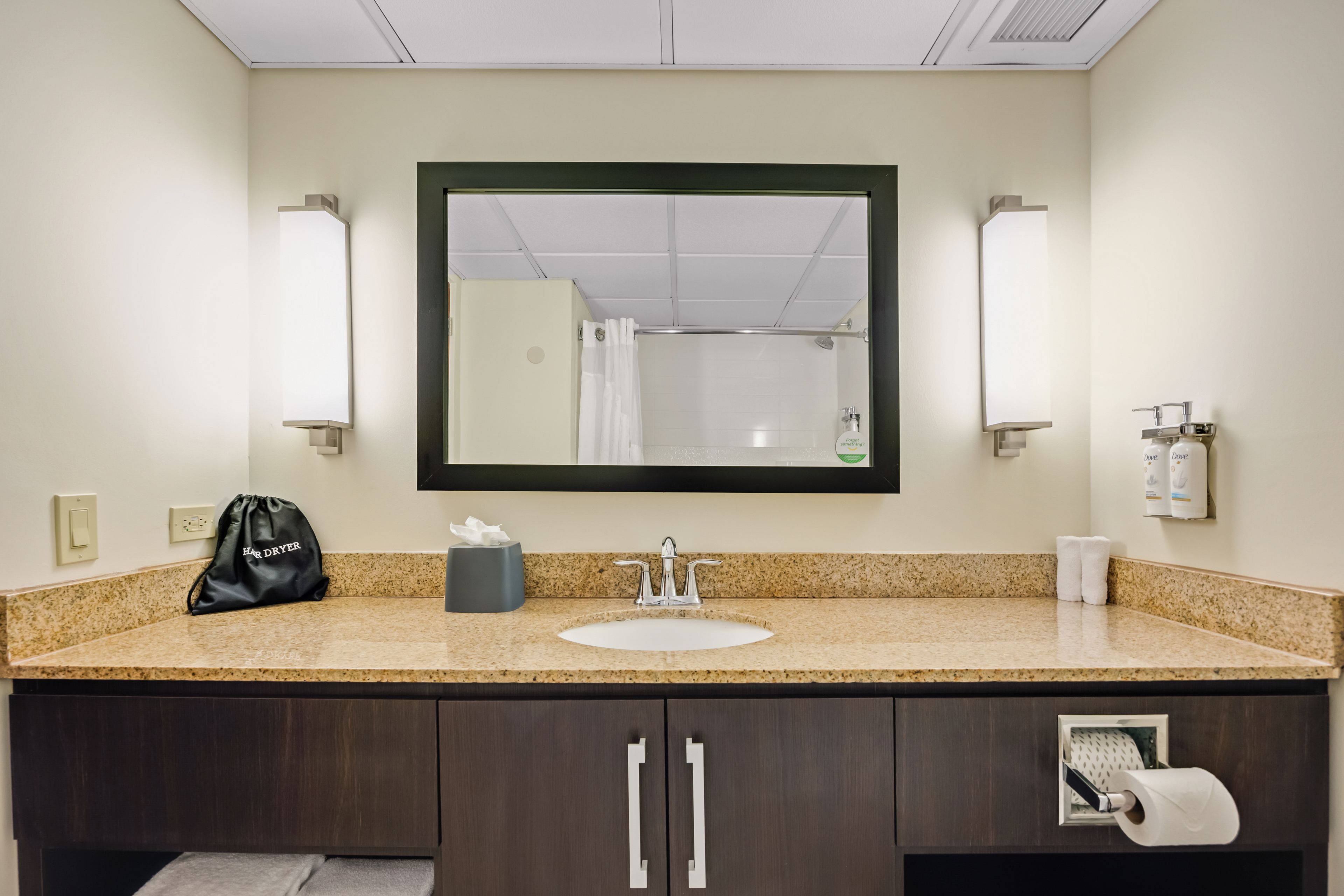 Baño de hotel con encimera de granito, lavamanos, espejo, toallero, secador de manos y dispensador de toallas de papel.