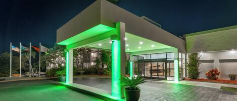 A modern hotel entrance with a covered walkway, illuminated by green lights, and a large potted plant at the entrance.