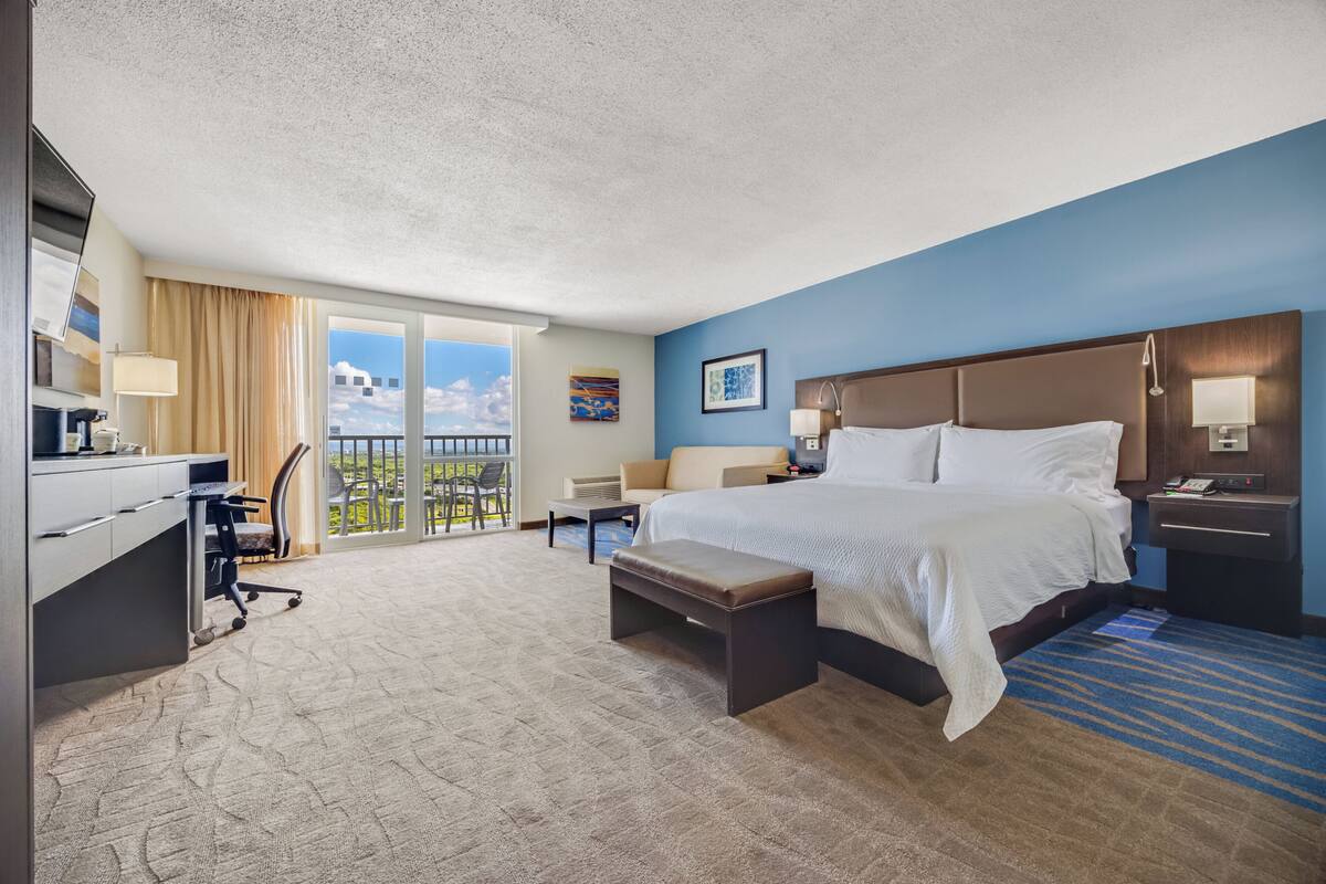A hotel room with a bed, desk, chair, and a balcony with a view.