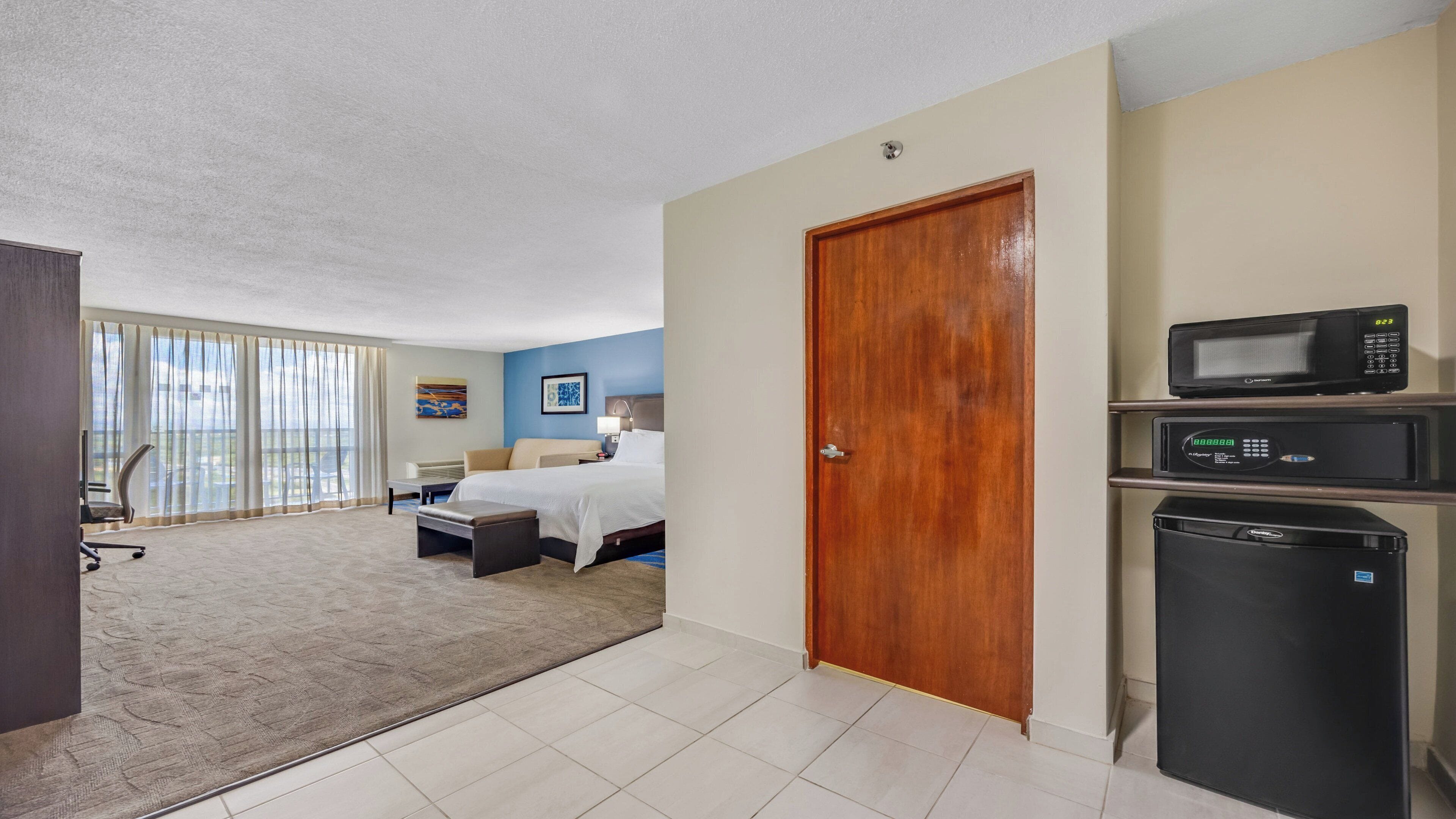 A hotel room with a bed, a desk, a chair, a microwave, and a refrigerator.