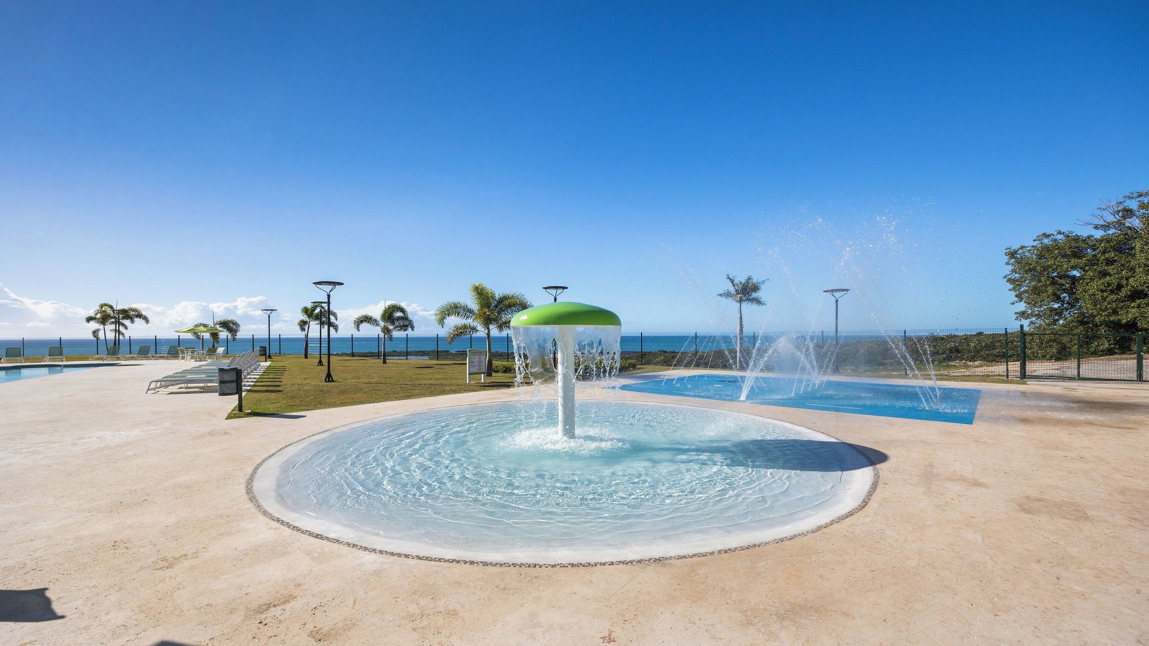 A circular water feature with a green top, surrounded by a pool and a beach view.