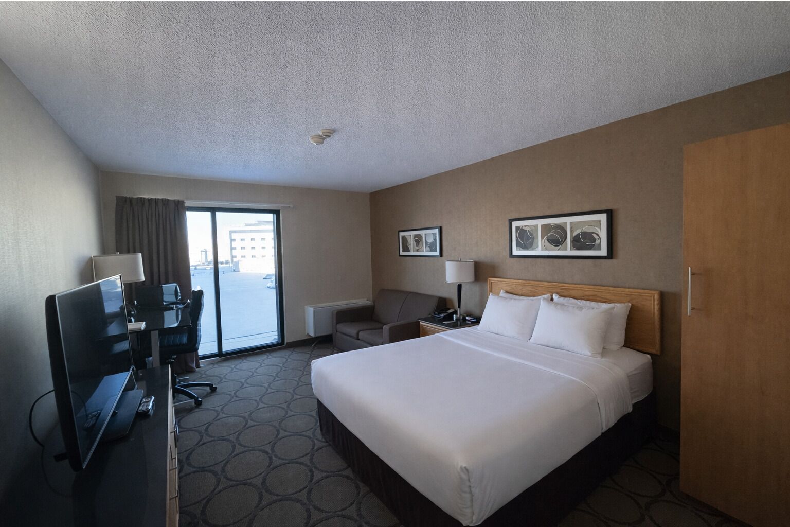 A hotel room with a large bed, a desk with a computer, a TV, and a view of the outside through a sliding door.