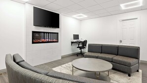 Une salle de conférence moderne, dotée d’un grand canapé d’angle gris, d’une table ronde, d’un téléviseur à écran plat fixé au mur et d’un bureau avec une chaise.