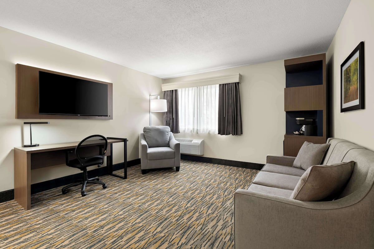 A hotel room with a flat-screen TV, a desk, a grey armchair, a sofa, and a patterned carpet.