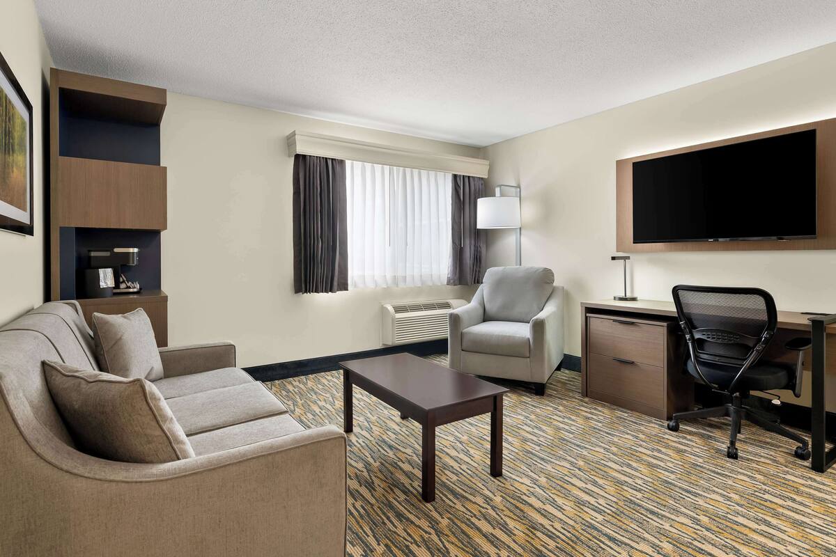 A hotel room with a sofa, armchair, desk, and a flat-screen TV.