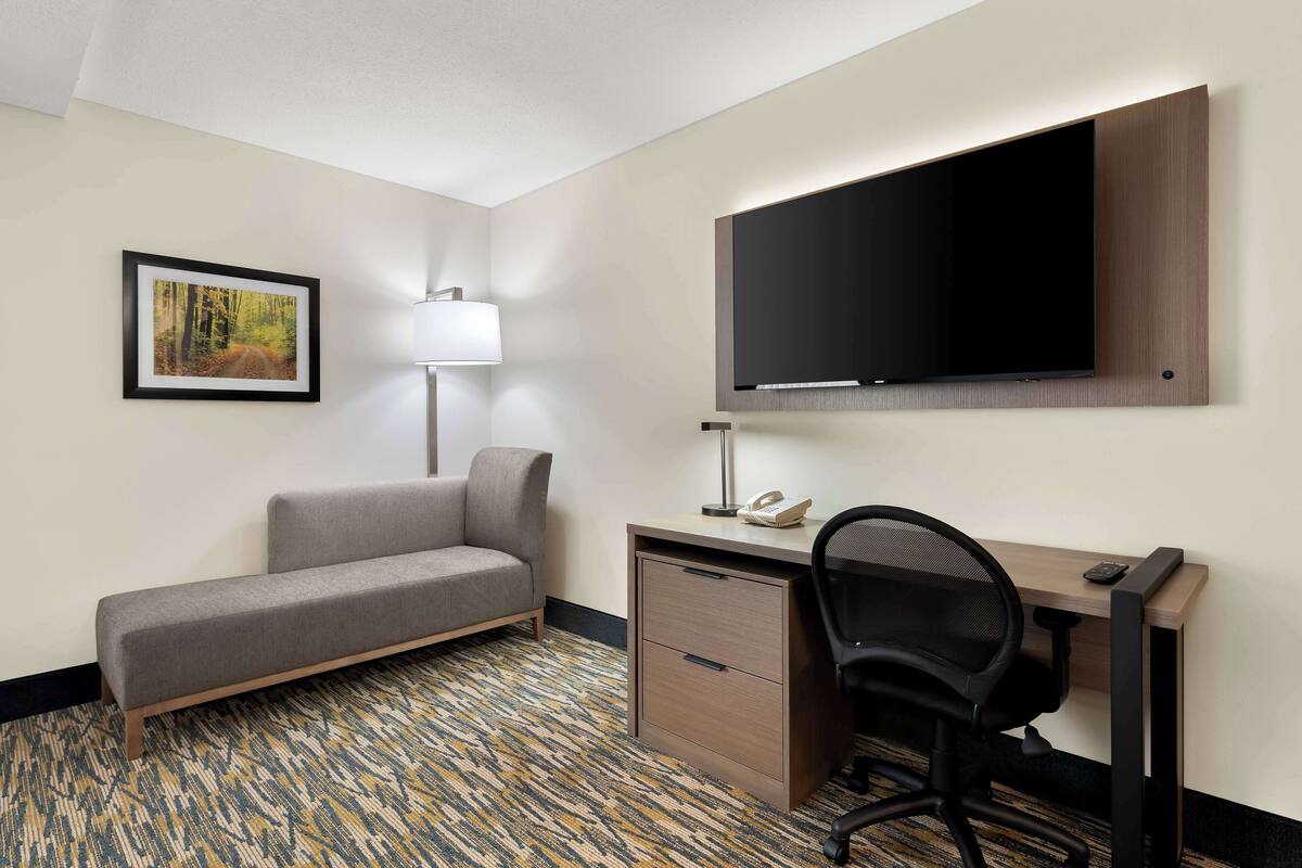 A hotel room with a sofa, desk, chair, flat-screen TV, lamp, and a framed picture on the wall.