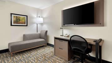 Ett hotellrum med en soffa, ett skrivbord, en stol, en platt-TV, en lampa och en inramad tavla på väggen.