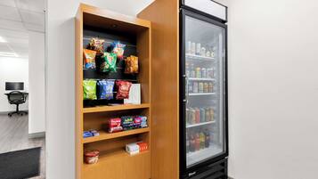 Máquina de vendas com snacks e bebidas, um frigorífico com bebidas e uma pequena cadeira de escritório ao fundo.