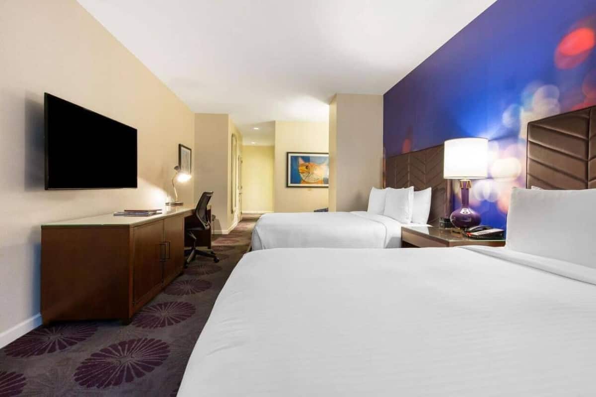 A hotel room with two beds, a flat-screen TV mounted on the wall, a desk with a lamp, and a painting on the wall.
