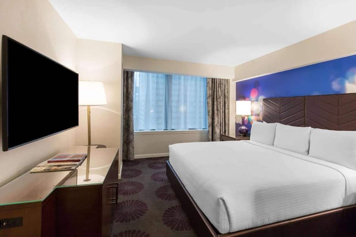 A hotel room with a large bed, a flat-screen TV, a desk with a lamp, and a window with curtains.