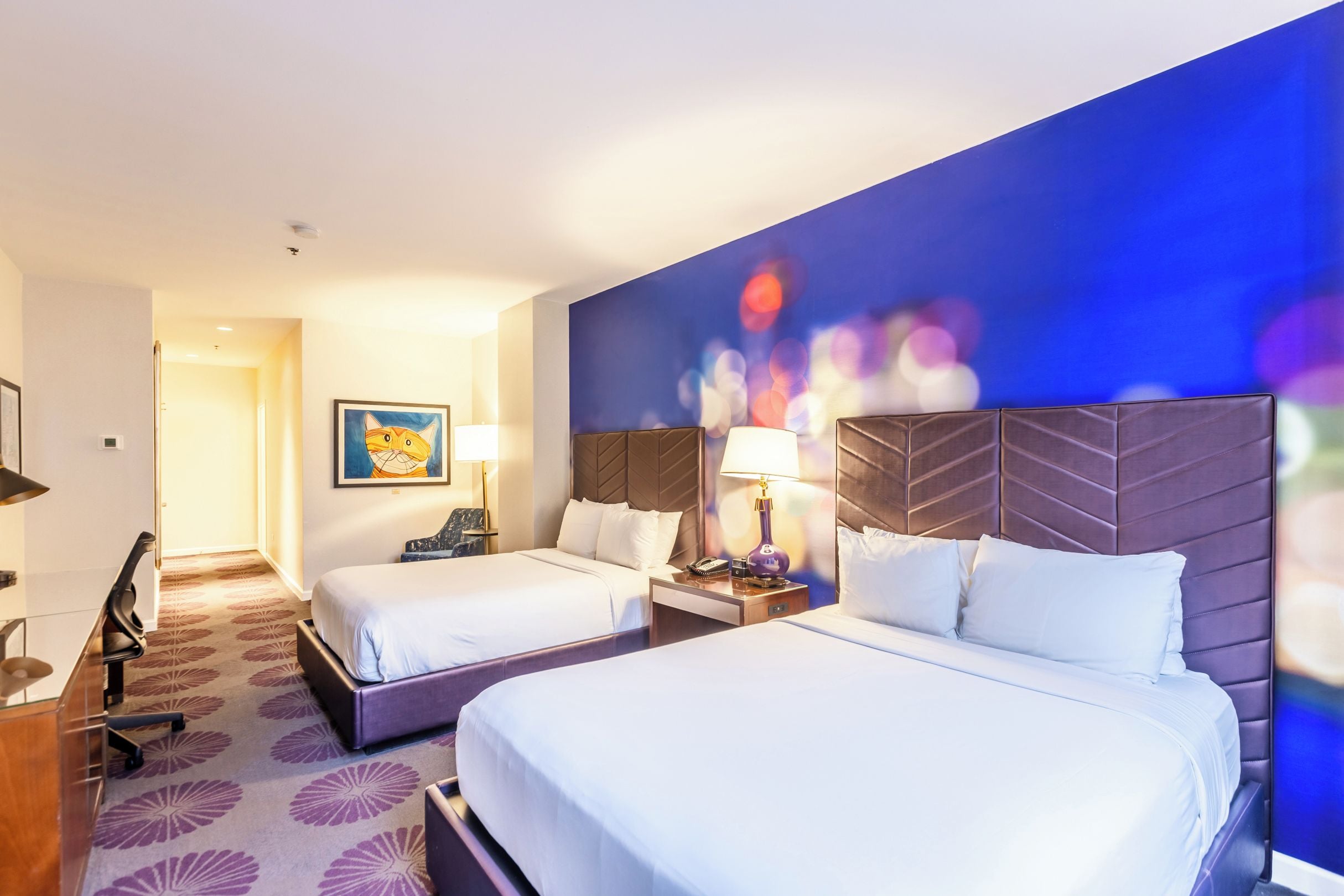 A hotel room with two beds, a desk, a chair, and a wall with a colorful abstract painting.