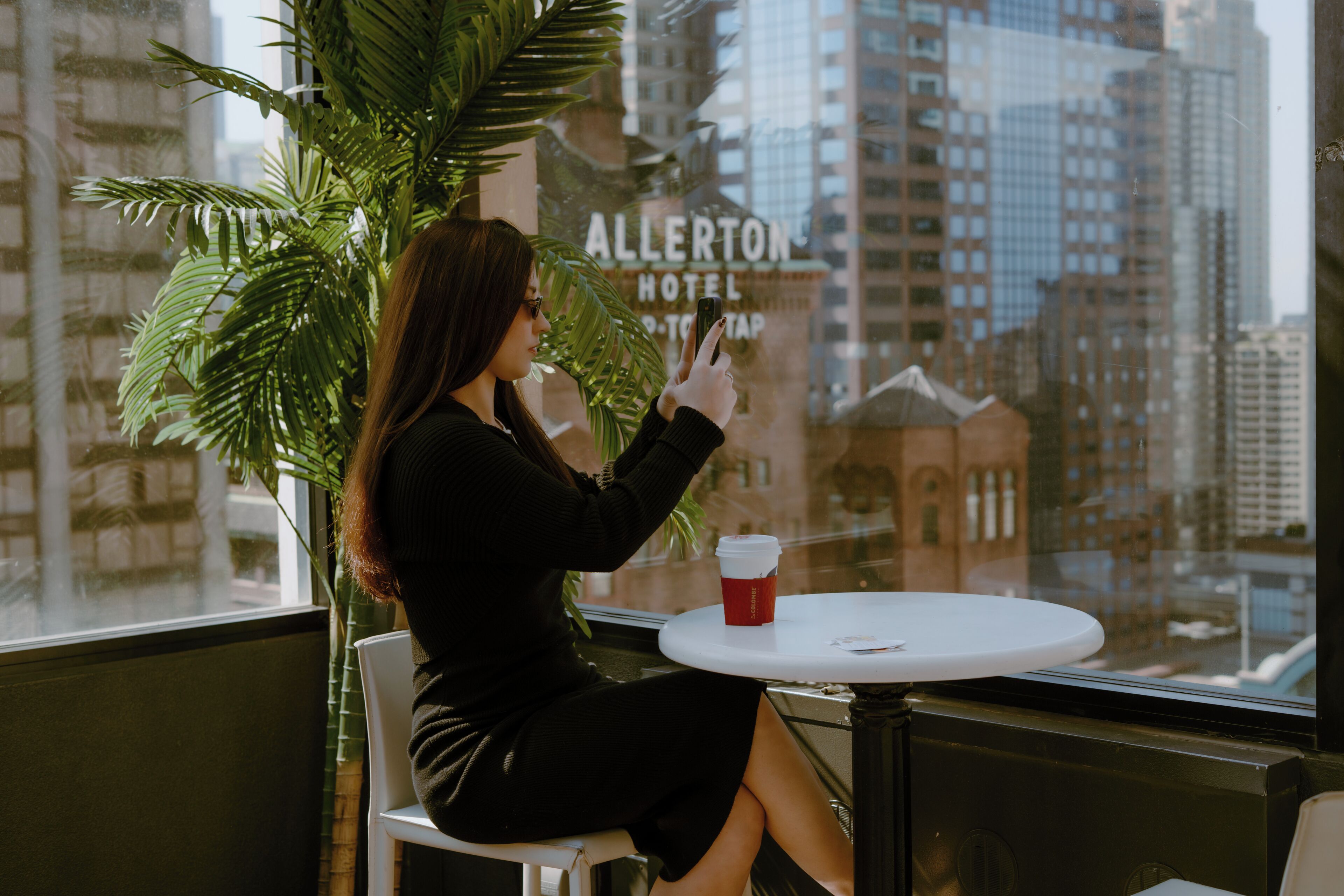 A person sitting at a table with a coffee cup, looking out a window at a cityscape with a building labeled "Allerton Hotel."
