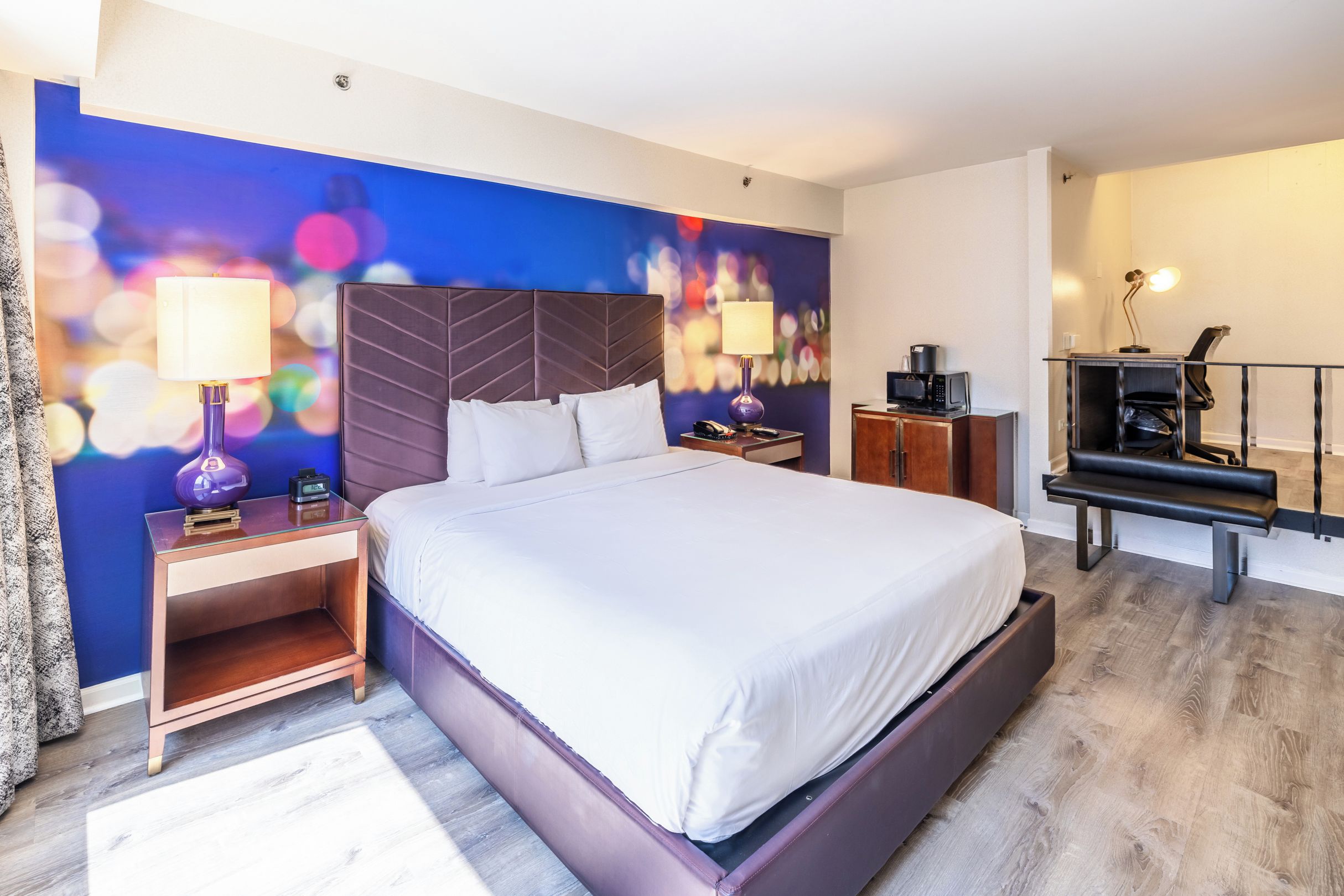 Une chambre d’hôtel avec un grand lit, un bureau avec une lampe, une petite kitchenette et un mur orné d’un motif abstrait et coloré.