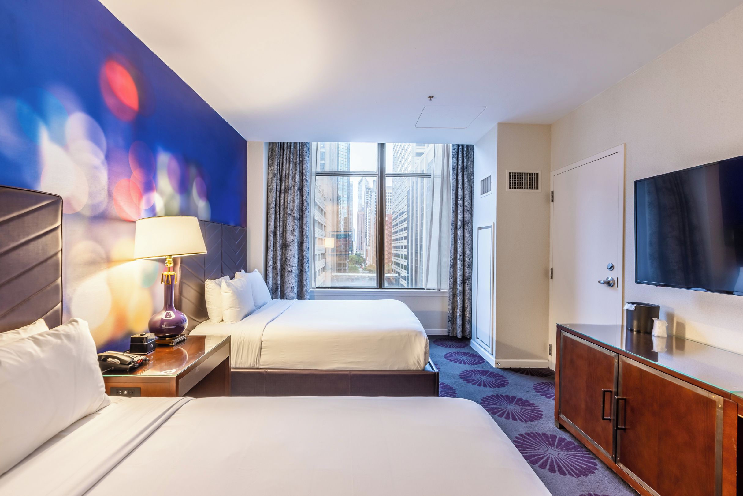 A hotel room with two beds, a TV, a desk with a lamp, and a window with a city view.