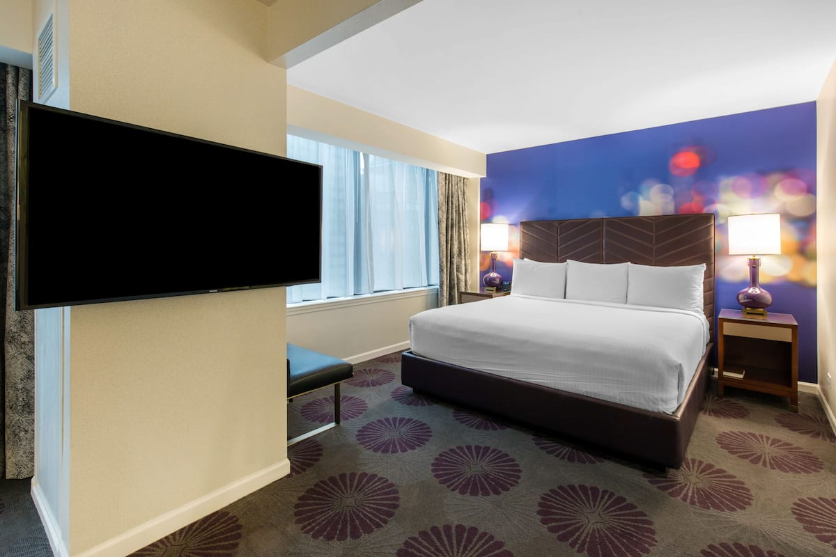 A hotel room with a large bed, a flat-screen TV mounted on the wall, a bedside table with a lamp, and a window with curtains.