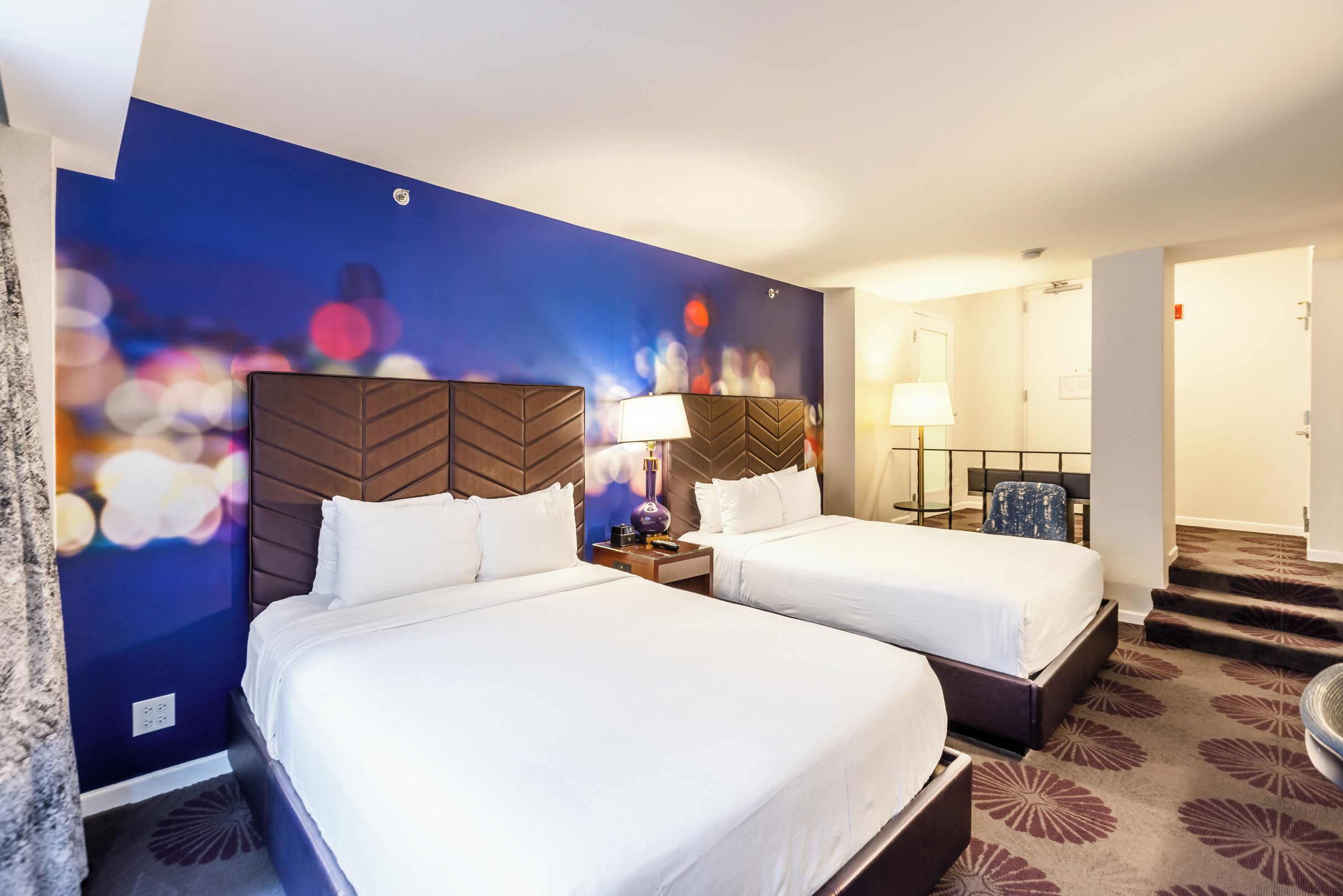Habitación de hotel con dos camas, una pared azul con efecto bokeh de luces y piso alfombrado.