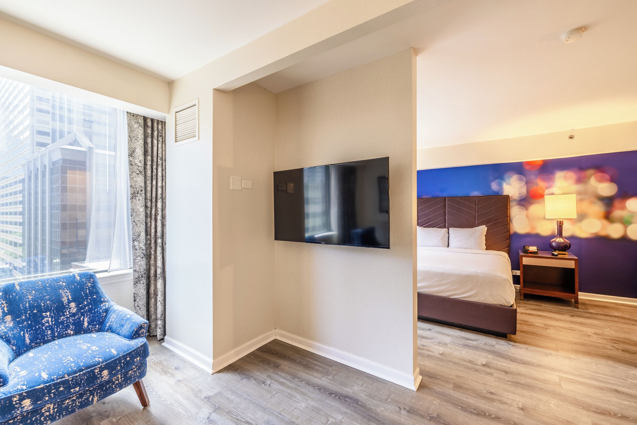 A hotel room with a large window, a flat-screen TV, a bed, a nightstand, and a blue armchair.