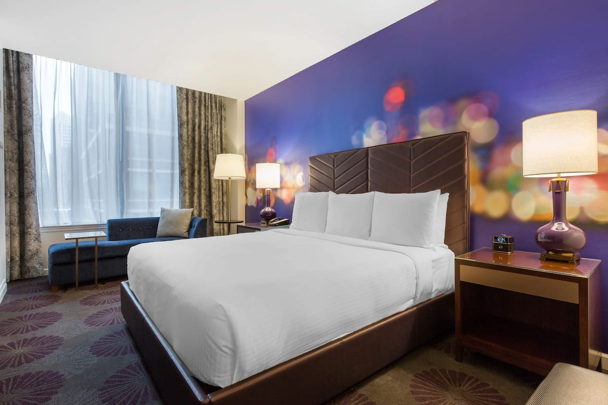 A hotel room with a large bed, a sofa, two nightstands with lamps, a window with curtains, and a colorful wall mural.